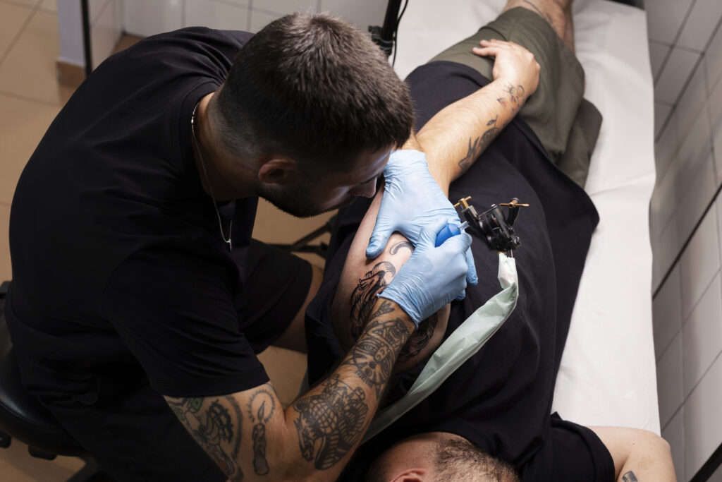 Un tatuador con camisa negra y guantes realiza un diseño detallado en la parte superior del brazo de una persona. El cliente se recuesta en una silla reclinable con un tatuaje parcialmente visible en el antebrazo. El entorno es un estudio de tatuajes muy iluminado.